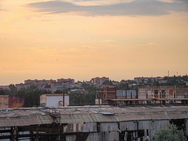 Görüntü, ön planda yıpranmış bir sanayi bölgesi ve arka planda alacakaranlık renkleriyle aydınlatılmış bir şehir silueti olan karanlık bir sahneyi gösteriyor.