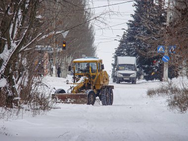 Gündüz vakti, yollarda kar temizleyen sarı bir buldozer, kısmen karanlık iki araç bir SUV ve bir dağıtım kamyonu, çıplak ağaçlar, bir Kostiantynivka, Ukrayna, 01-15-2013