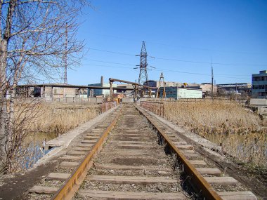 Terk edilmiş bir demiryolu hattının görüntüsü, her iki tarafında bitki örtüsü ve arka planda endüstriyel altyapı var. Gökyüzü açık. Kostiantynivka, Ukrayna, 06-15-2013