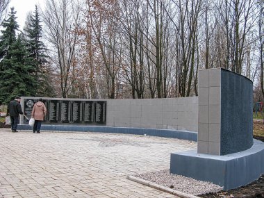 Görüntü, önünde iki ziyaretçinin durduğu yapraksız ağaçlarla çevrili bir açık taş anıtı tasvir ediyor. Yapının görünür metinleri olmayan plaketleri var ve Kostiantynivka, Ukrayna, 06-15-2013