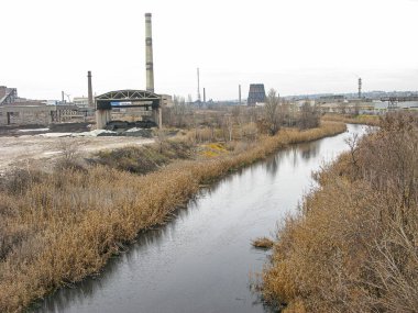 Bulutlu bir görüntü, bir su yolu, muhtemelen bir kanal ya da nehir olan bir sanayi bölgesini gösteriyor. Su kütlesi kurak bitki örtüsüyle çevrelenmiş, bu da yaz sonu veya Kostiantynivka, Ukrayna, 06-15-2013
