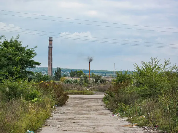 Bulutlu bir gökyüzünün altında terk edilmiş bir sanayi bölgesi, eski bir bina veya tesise doğru uzanan beton bir yol ile uzaktaki bacalar, borular Kostiantynivka, Ukrayna, 06-15-2013