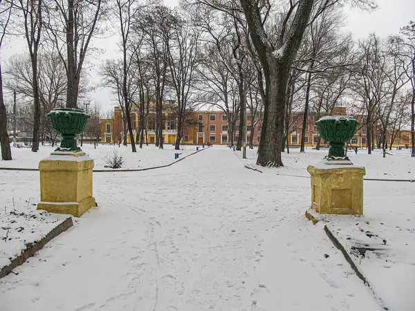 Şehir parkından geçen karla kaplı patikalı bir kış sahnesinde, önde çıplak ağaçlar, iki dekoratif vazo ve Kostiantynivka, Ukrayna 'da geleneksel mimari, 01-15-2013