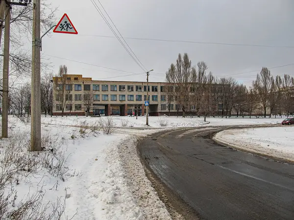 Karlı bir şehir manzarası, kısmen temizlenmiş bir yol, sağda endüstriyel veya eski bir konut binası, solda bir trafik uyarı levhası, ve Kostiantynivka, Ukrayna 'da çıplak ağaçlar, 01-15-2013