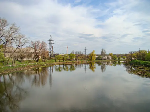 Şehir parkında yeşil ağaçlar, bulutlu gökyüzünü yansıtan sakin sular ve arka plandaki Kostiantynivka, Ukrayna 'daki sanayi yapıları arasında geçen sakin bir nehir sahnesi, 01-15-2013