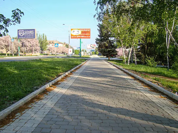 Çimlerle kaplanmış ve ağaçlarla gölgelenmiş, kentsel alana doğru uzanan kaldırımlı bir yürüyüş yolu sol tarafında okunamayan işaretlerle hafif eğimli Th Kostiantynivka, Ukrayna, 06-15-2013