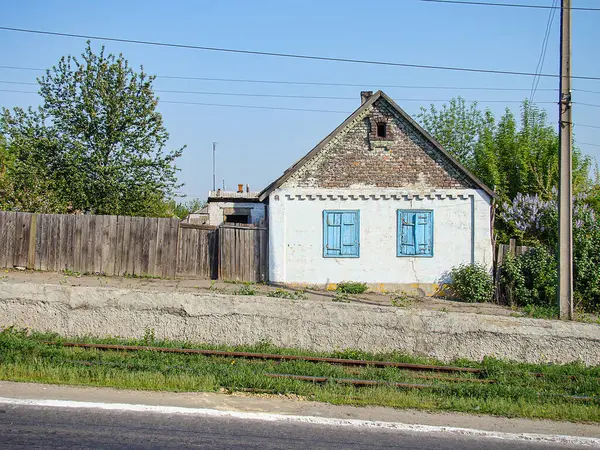 Küçük, tek katlı, gri dış ve beyaz yamalı kır evi, mavi panjurlu iki pencere, merkez kapı, düz olmayan zemin, toprak yol, ahşap fen Kostiantynivka, Ukrayna, 06-15-2013