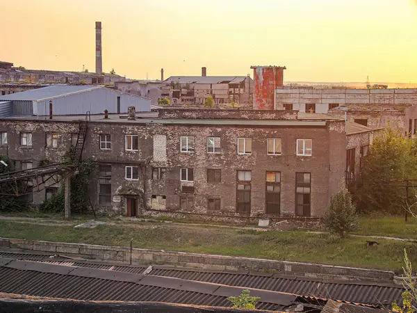 Terk edilmiş bir sanayi binası, muhtemelen 19. yüzyılın sonlarından ya da 20. yüzyılın başlarına ait, turuncu ve sarı günbatımlı bir gökyüzünün zeminine karşı resmedilmiştir.