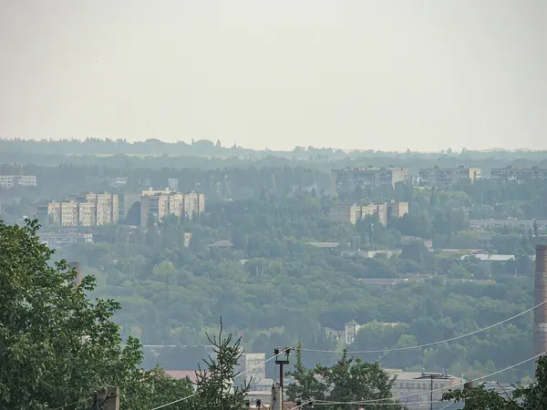 Şehir manzarası yüksek bir perspektiften, binaların karışımı, bulutlu gökyüzü ve önplanda bir ağaç kümesi ile görünüyor. Tarz gerçekçi Kostiantynivka, Ukrayna, 06-15-2013
