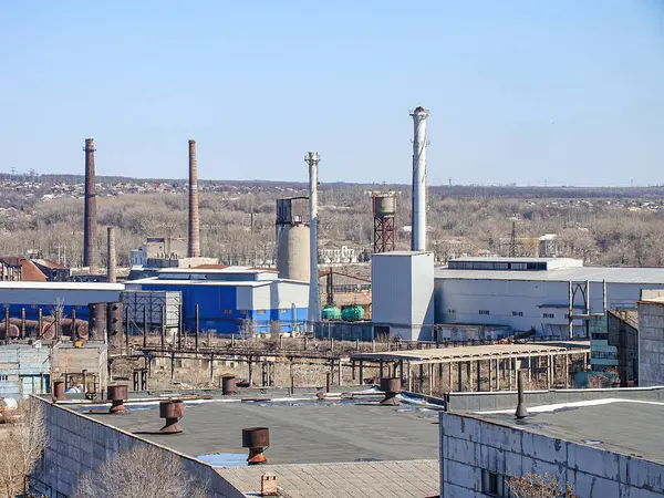 Görüntü, açık bir gökyüzü altında birden fazla bina, baca ve havalandırma kanalları bulunan bir endüstriyel kompleksi gösteriyor. Kompleks, seyrek, çıplak dallı Kostiantynivka, Ukrayna, 06-15-2013