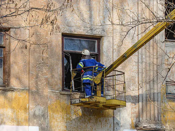 Koruyucu giysili bir işçi kovanın içinden vinç kullanarak eski ve hasarlı bir binayı onarıyor. Sahne çıplak sonbahar ağaçları ve Kostiantynivka, Ukrayna, 01-15-2013