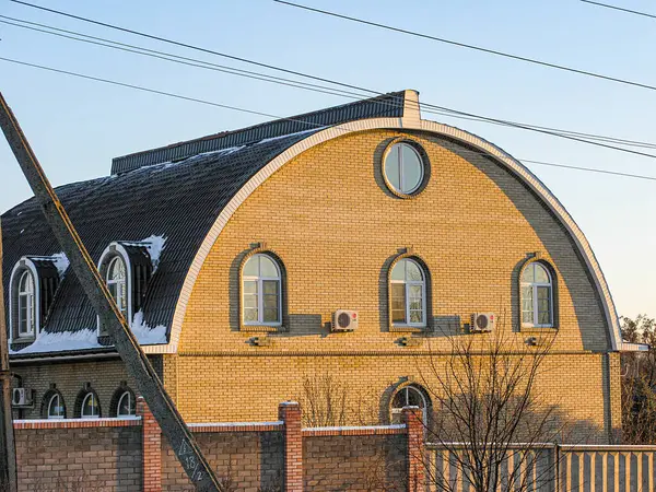 Görüntü, turuncu ve sarı günbatımı gökyüzüne karşı uzanan eğri çatılı eşsiz bir tuğla bina gösteriyor. Binanın yuvarlak pencereleri var, kısmen tahta Kostiantynivka, Ukrayna, 01-15-2013