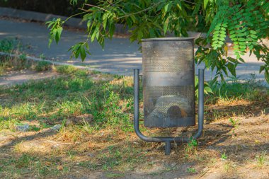 Çimlerin üzerindeki metal silindirik ateş çukurunun açık hava görüntüsü, muhtemelen ağaç örtüsü olan parka benzer bir ortamda ve görülebilir insan yok.