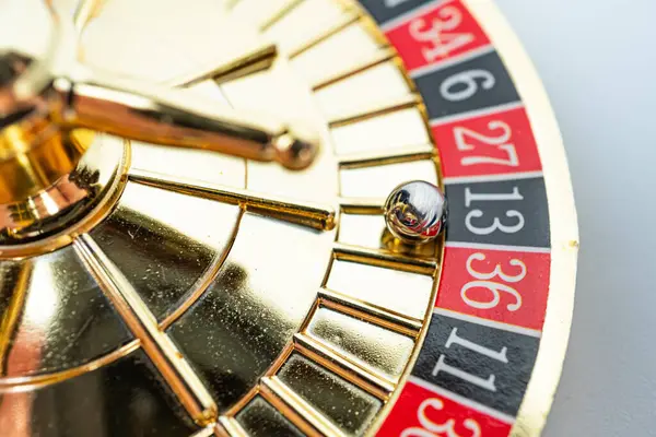 Rulet tekerleğinin yakın plan çekimi, numaralandırılmış bölmeleri ve raydaki metalik topu gösteriyor. Tasarım altın arka planda kırmızı ve siyah sayılar içeriyor, lüks ve oyun temasını çağrıştırıyor.