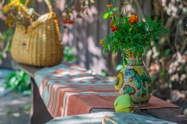 Kırsal unsurlar, dokuma masası, canlı çiçeklerden oluşan bir vazo ve geniş dokuma sepetinin yer aldığı açık hava ortamında, çevre dostu ürün pazarlaması için ideal, bereketli bir bahçe görülüyor.