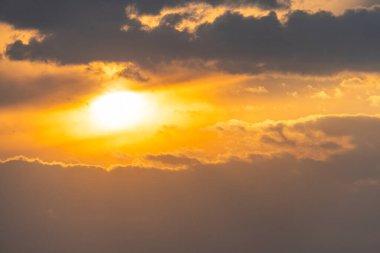 Bir resim, güneşin kısmen bulutlar tarafından gizlenmiş, turuncudan altına dönüşen, dinginliği, doğal güzelliği veya yeni başlangıçları yansıtan dramatik bir gökyüzünü gösteriyor.