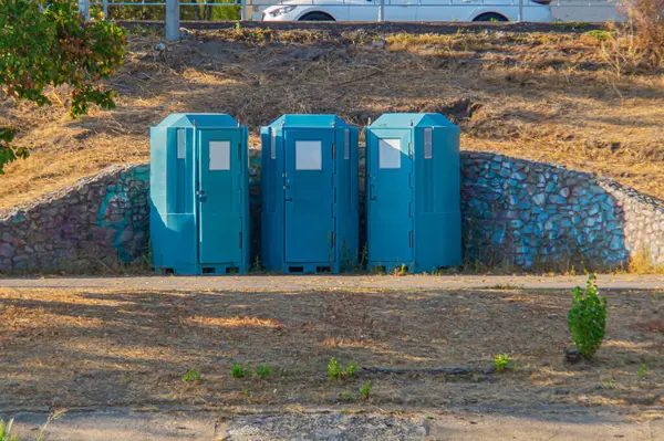 Üçgen biçiminde üç mavi portatif tuvaletli açık hava sahnesi, kayalık bir zemin, sağlık hizmetleri, festivaller, etkinlikler veya temizlik pazarlama için kullanılan engebeli bir ortamı işaret ediyor.