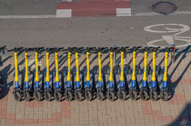 Görüntü, şehir ortamında kaldırımda bir sıra mavi elektrikli scooter gösteriyor, muhtemelen çevre dostu şehir ulaşımını desteklemek için kullanılan bir kiralama şirketinden, Kyiv, Ukrayna, 09-15-2024
