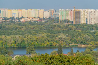 Doğayla kalkınma arasındaki uyumu sembolize eden yemyeşil ve durgun su zeminine karşı çeşitli mimari tarzlara sahip kentsel ufuk çizgisi Kyiv, Ukrayna, 09-15-2024