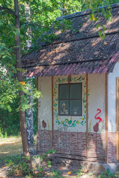 Ormanlık bir alanda sazdan bir çatısı olan küçük, kırsal ahşap bir kulübe, çiçek desenleri ve dekoratif pencere çerçevesi sükunet ve sadelik yayıyor, ideal Kyiv, Ukrayna, 09-15-2024