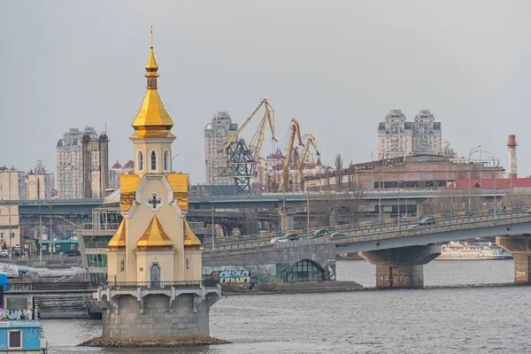 Altın kubbeli bir kilise ve zıt sarı bir binanın yer aldığı şehir sahnesi, tarihi ihtişamı ve modernliği sembolize ediyor. Sakin su vibra Kyiv, Ukrayna, 09-15-2024 'ü yansıtıyor.