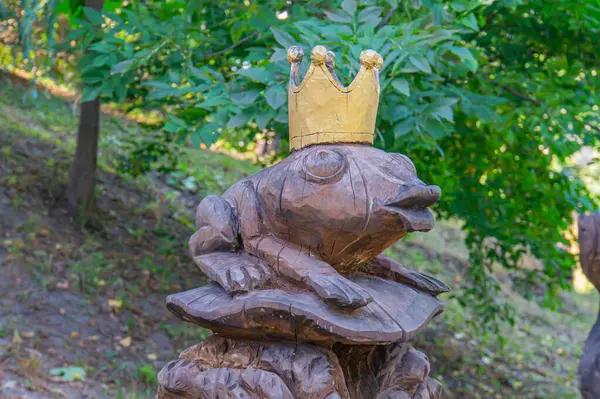 Antropomorfik bir kurbağanın açık hava heykeli altın bir taç takıyor, diğer oyma figürlerle çevrili doğal bir çevrede, tuhaf ve fantastik tarzda, muhtemelen bir Kyiv, Ukrayna, 09-15-2024