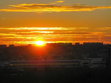 Yüksek bir bakış açısından nefes kesici bir gün batımı manzarası, binalar ve bacalarla dolu kentsel bir manzara, hepsi sıcak, canlı renklerle yıkanmış.