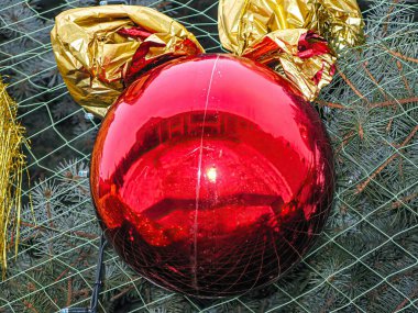 Yansıtıcı yüzeyi olan parlak kırmızı bir Noel süsü bir Noel ağacında asılı, kısmen altın sarısı bir nesne tarafından gizleniyor.