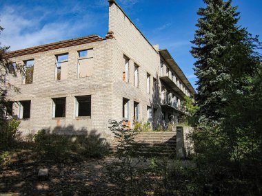 İhmal edilmiş çok katlı tuğlalı bir bina, açık tuğlalar, soyulmuş boyalar, kayıp pencere çerçeveleri ve çevredeki aşırı gelişmiş bitki örtüsü. Görüntü açık bir günde çekilmiş.