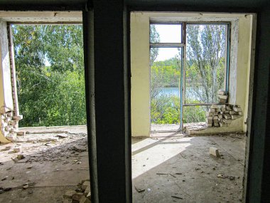 Açık duvarları ve enkazı olan boş, dağınık bir oda pencerelerden geçen gün ışığı açık yeşilliği ortaya çıkarıyor, bitki örtüsü yakınındaki bir yeri gösteriyor.