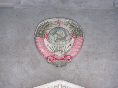 Bir çember içinde çekiç ve orak amblemi resmi, muhtemelen komünist bir rejimle ilişkili, dokulu gri bir arka planda ek dekoratif unsurlar ve mimari özellikler görünür ab
