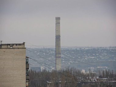 Görüntü, gri gökyüzüne duman yayan büyük bir baca ile endüstriyel enerji santrali sahnesini gösteriyor. Çok katlı bina, görünür pencereler ve kontrolle betondan veya benzer malzemelerden oluşuyor.