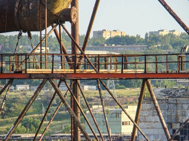 Görüntü, bitki örtüsü olan terk edilmiş bir sanayi yapısını, ön planda daha küçük yapıları, uzaktaki konut binalarını gösteriyor.