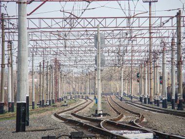 Çoklu rayları olan bir elektrikli demiryolu deposu, tepedeki katenareri yapılar, güvenlik bariyerleri ve bulutlu gökyüzünün altında trafik ışıkları