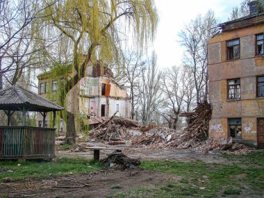 Görüntü, muhtemelen Sovyet sonrası terk edilmiş bir şehir manzarasını gösteriyor. Yıkık bir bina, aşırı gelişmiş yeşillikler, çimenler, çıplak toprak, molozlar, yapraksız ağaçlar, kasvetli bir atmosfer, açık hava koltuğu.