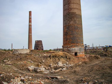 Endüstriyel bozulma sahnesi, bozulan iki yapı, bitkisel büyüme, enkaz ve seyrek çimen ile dağılmış düzensiz zemin veya arka planda yosun puslu gökyüzü