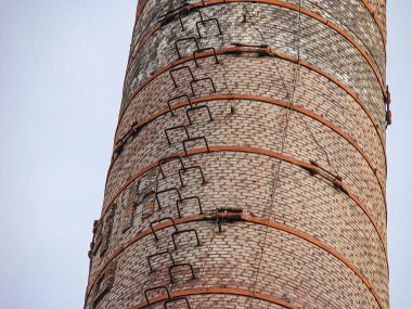 Görüntü uzun, tuğla ya da metal bacayı gösteriyor. Çoklu katmanlı ve dikey çubuklu. Gökyüzü bulutlu ve düzensiz aydınlatma, görünmeyen yapıların gölgelerini gösteriyor.