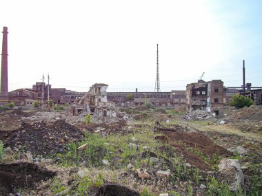 Endüstriyel dönüşüm sonrası terk edilmiş bir sanayi manzarası ya da çevresel restorasyon enkazı, bitkiler ve molozlar, harabelerin arasında duran uzun bir baca ile bölgeye hükmediyor.