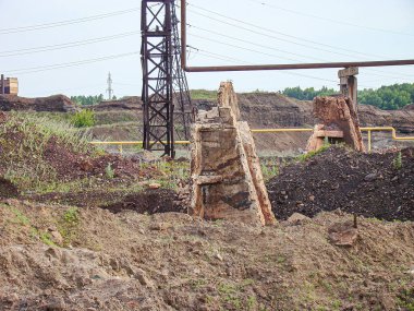 Merkezde yıpranmış beton ya da taş bir sütun, yakınlarda kazılmış malzemeler ve bulutlu gökyüzünde çaprazlama çalışan elektrik hatlarının yer aldığı endüstriyel bir manzara.
