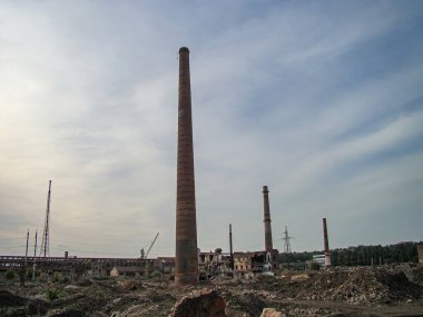 Harap olmuş bir baca ve diğer binaların bulunduğu terk edilmiş bir sanayi bölgesi. Bitki örtüsü ve enkazla kaplanmış. Olay yeri parçalı bulutlu.