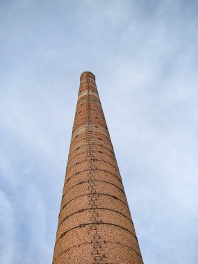 Bulutlu bir gökyüzüne karşı eski, kırmızı tuğla bir baca. Yapısı silindirik ve tepede daralıyor.