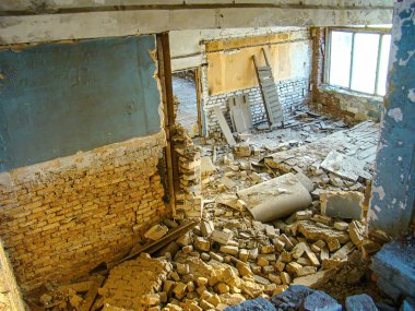 Görüntü, yıkıntılarla, yıkılan parçalarla ve açıkta kalan tuğlalarla dolu terk edilmiş ya da güvensiz bir binanın hasarlı iç kısımlarını gösteriyor. Tavanda görünür çatlaklar var.