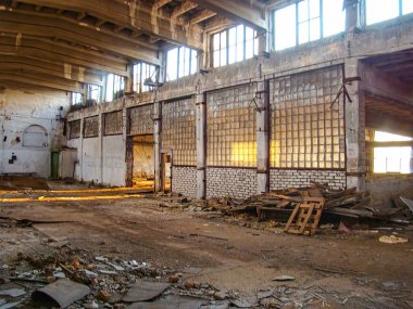 Düz zemini, enkazı, açıkta kalan kirişleri, tavan pencereleri, paslanmış metal panelleri, büyük kapıları olan harap bir depo.