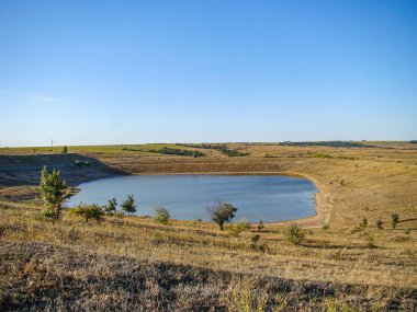 Çorak zemin, seyrek bitki örtüsü, durgun göl, yumuşak yamaç yavaş dağlar, parçalı bulutlu gökyüzü, öğleden sonra aydınlatması, görünür insan veya metin yok.