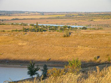 Doğal bir manzaranın panoramik bir görüntüsü, kuru otlaklar, yeşil bitki örtüleri, bir su kütlesi nehri veya göl, yuvarlanan tepeler, ve berrak mavi bir gökyüzü.