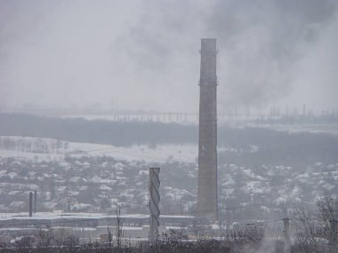 Kış günü, karla kaplı ağaçlar ve yapılarla kaplı endüstriyel bir manzara.