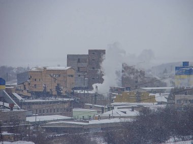 Karlı bir şehir manzarası, büyük, endüstriyel binalar, dalgalanan bacalar, ve canlı sarı-mavi bir bina gökyüzü gridir ve genel renk paletine beyaz ve gri gölgeler hakim.