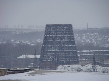 Belirgin bir kuleye ve çeşitli makinelere sahip karlı arazinin ortasındaki büyük bir endüstriyel yapının görüntüsü durağan sahneye hareket katıyor, ön planda küçük binalar görünüyor.
