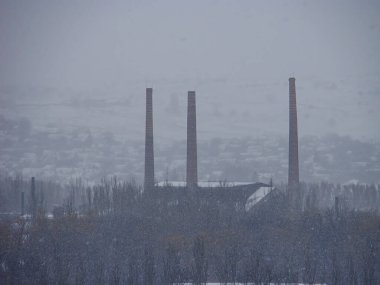 Görüntü, kış manzarasında üç ince duman bacası bulunan karlı bir sahneyi gösteriyor. Gramsi dokular, muhtemelen eski bir fotoğraf veya değiştirilmiş. Görünür bir metin içeriği yok.