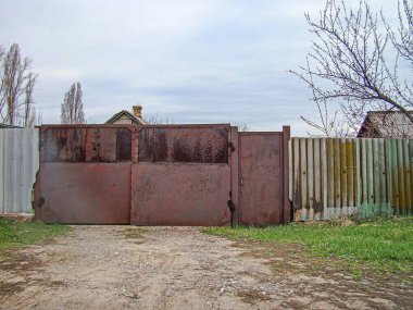 Eski paslı metal kapı, yatay çıtçıtlı ve yatay takviye çubuklu. Açık bir alanda. Her iki tarafta seyrek bitki örtüsü ya da çimen var.